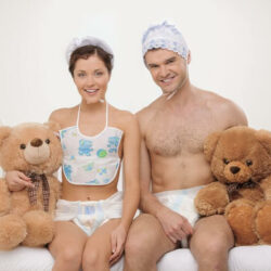 A woman and a man dressed as babies sit on a crib with teddy bears at their sides.