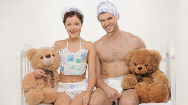 A woman and a man dressed as babies sit on a crib with teddy bears at their sides. 