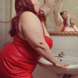 A woman with long red hair dressed in a sexy red dress posing in front of a sink and gold mirror. 
