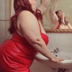 A woman with long red hair dressed in a sexy red dress posing in front of a sink and gold mirror. 