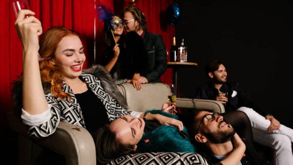 A group of smiling people at a party sitting close, reclining on a couch and on each other's laps. 