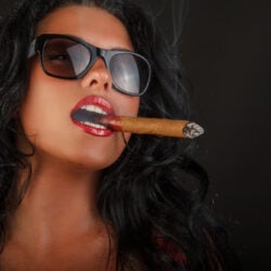 Hot woman with long dark hair, sunglasses and red lipstick smokes a cigar.