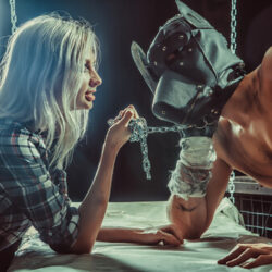 A blonde woman in a flannel shirt pulls a leather masked man closer using an attached chain.