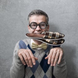 A man in an argyle sweater, bow tie and glasses poses like a puppy with plaid slippers in his mouth. 