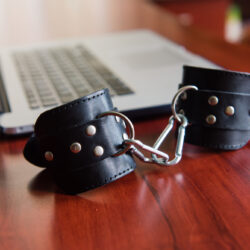 Leather studded handcuffs sitting on a polished cherry wood desk next to an open laptop. 
