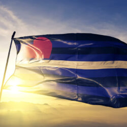 A kink flag blows in the wind in front of a blue sky with the sun behind it. 