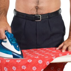 A shirtless man wearing business pants and belt ironing a red floral fabric.