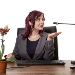 A boss lady in dressed in a suit sitting behind a desk outstretches her palm to accept a kinky toy.