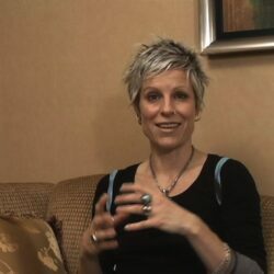 A woman with short, spiky, platinum blonde hair, a black shirt with three quarter length sleeves sits on a brown couch talking into the camera and gesturing with her hands.