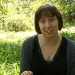 A woman with dark hair in a chin length bob and bangs is sitting in a grassy wooded area.  She is wearing a black scoop neck shirt and cardigan. She is talking into a camera.