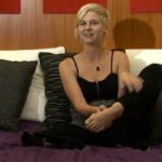 A woman with short pixie style blonde hair is sitting on a bed facing the camera.  She is wearing a black tank top and black pants.  Her left foot is raised a little to show the camera.