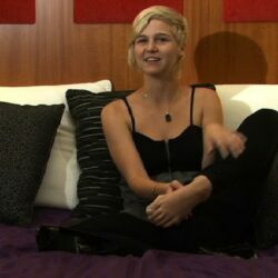 A woman with short pixie style blonde hair is sitting on a bed facing the camera.  She is wearing a black tank top and black pants.  Her left foot is raised a little to show the camera.