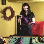 A woman with long black hair, blunt bangs, and wearing a short-sleeve black shirt is standing behind a table covered with floggers. She is holding one flogger stretched between her hands.