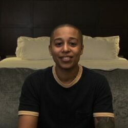 A person with closely shaved head, wearing a black short sleeve shirt with tan trim is sitting on the end of a bed and speaking into the camera.