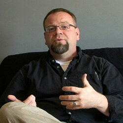 A man with very short hair, glasses, goatee, and wearing a dark button down shirt is sitting on a couch and speaking to the camera.