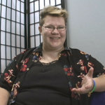 A woman with short spiky blonde hair, glasses and wearing a black and floral collared shirt open over a black shirt is sitting and speaking into the camera.