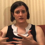 A  woman with chin length dark hair, wearing a sleeveless black top with white neckline, is sitting and talking into the camera.