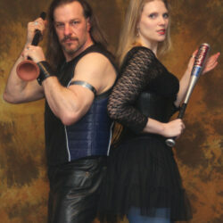 A man with long hair, a goatee, and wearing leather pants and leather vest is standing back to back with a woman.  She has long blonde hair, blue leggings and a black frilly mini dress.  He is holding a plunger in both hands and she has a small metal bat.