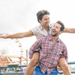 A man happily getting a piggy back ride from another man with arms out and a ferris wheel in the background.