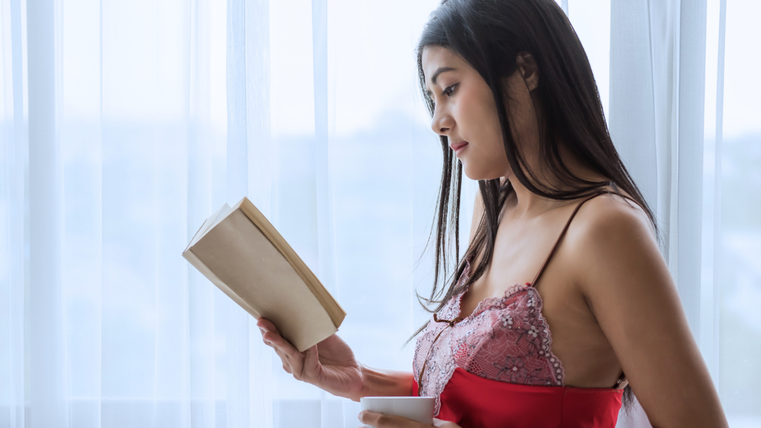Photo of a feminine person in red lingerie reading a book in front of a window