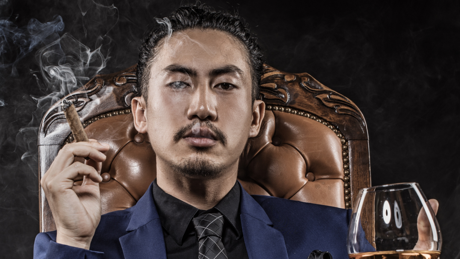 A masculine Asian person wearing a suit and smoking a cigar in a brown leather chair