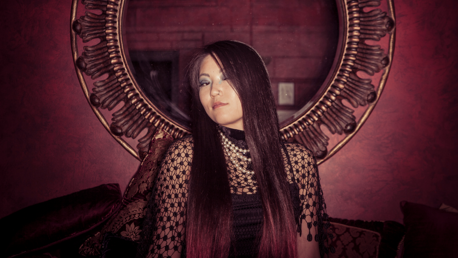 A feminine Asian person with long hair has a confident expression and sits in front of a round wall mirror