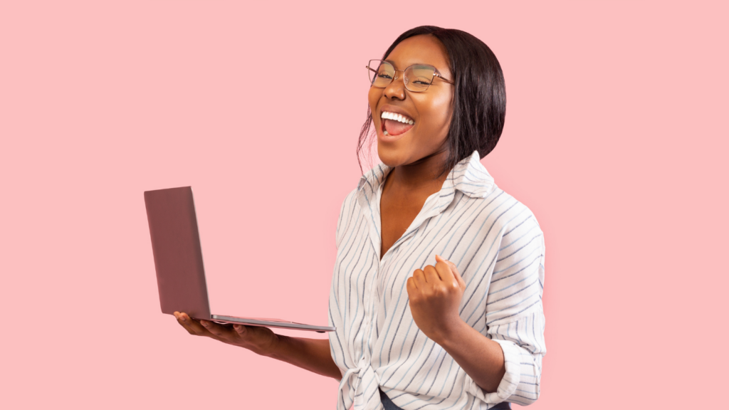 Photo of a feminine person with brown skin and metal frame glasses holding up a laptop and looking excited