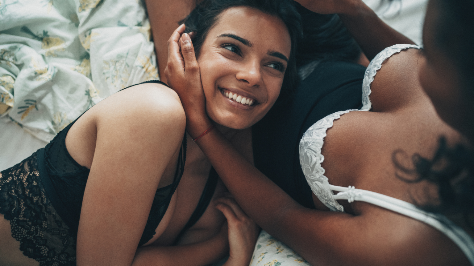 Photo of two feminine looking partners, both with brown skin, one wearing white lingerie and the other wearing black lingerie. One parter has their head in the other's lap and is smiling up at them