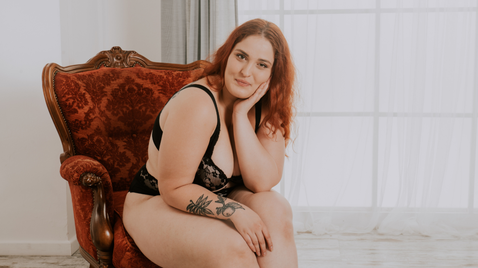 Photo of a plus size feminine person with light skin and red hair and tattoos on their arm sitting on a velvet chair and smiling softly