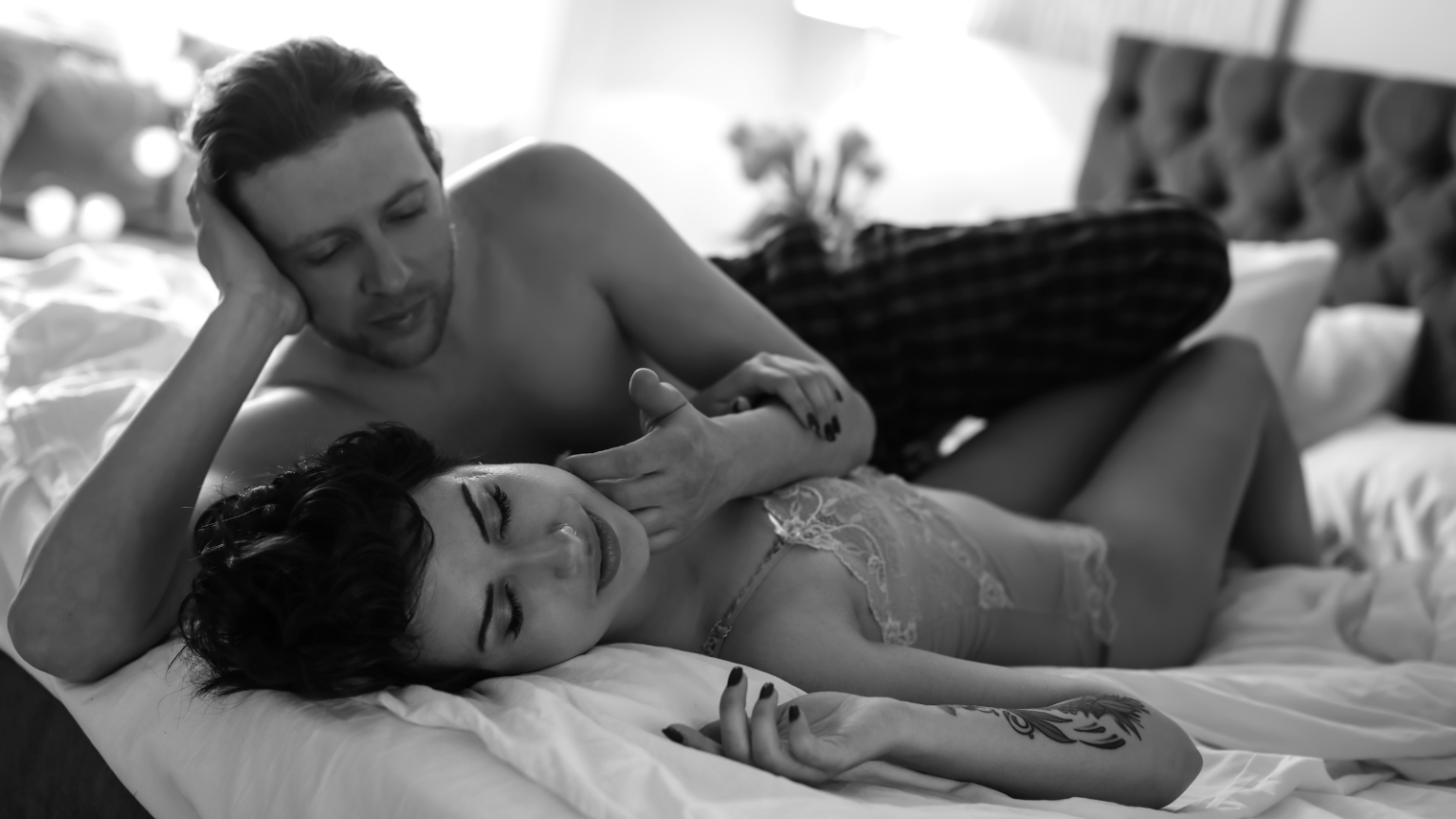 Two partners lying in bed, one masculine looking and the other feminine looking, both with light skin. The masculine partner is gently stroking the face of the feminine partner