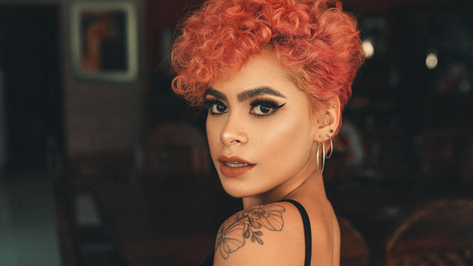 Photo of a feminine looking person with light brown skin and dyed red curly hair looking at the camera with a relaxed but serious expression