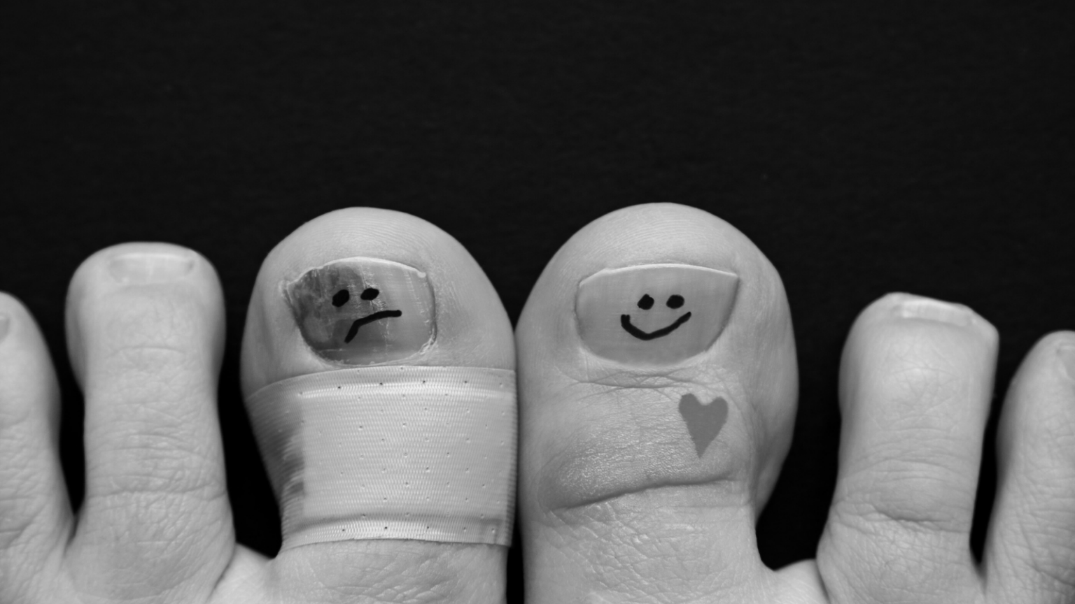 Closeup photo of toes. One has a band aid and a sad face drawn on the toenail. A happy face is drawn on another toenail.