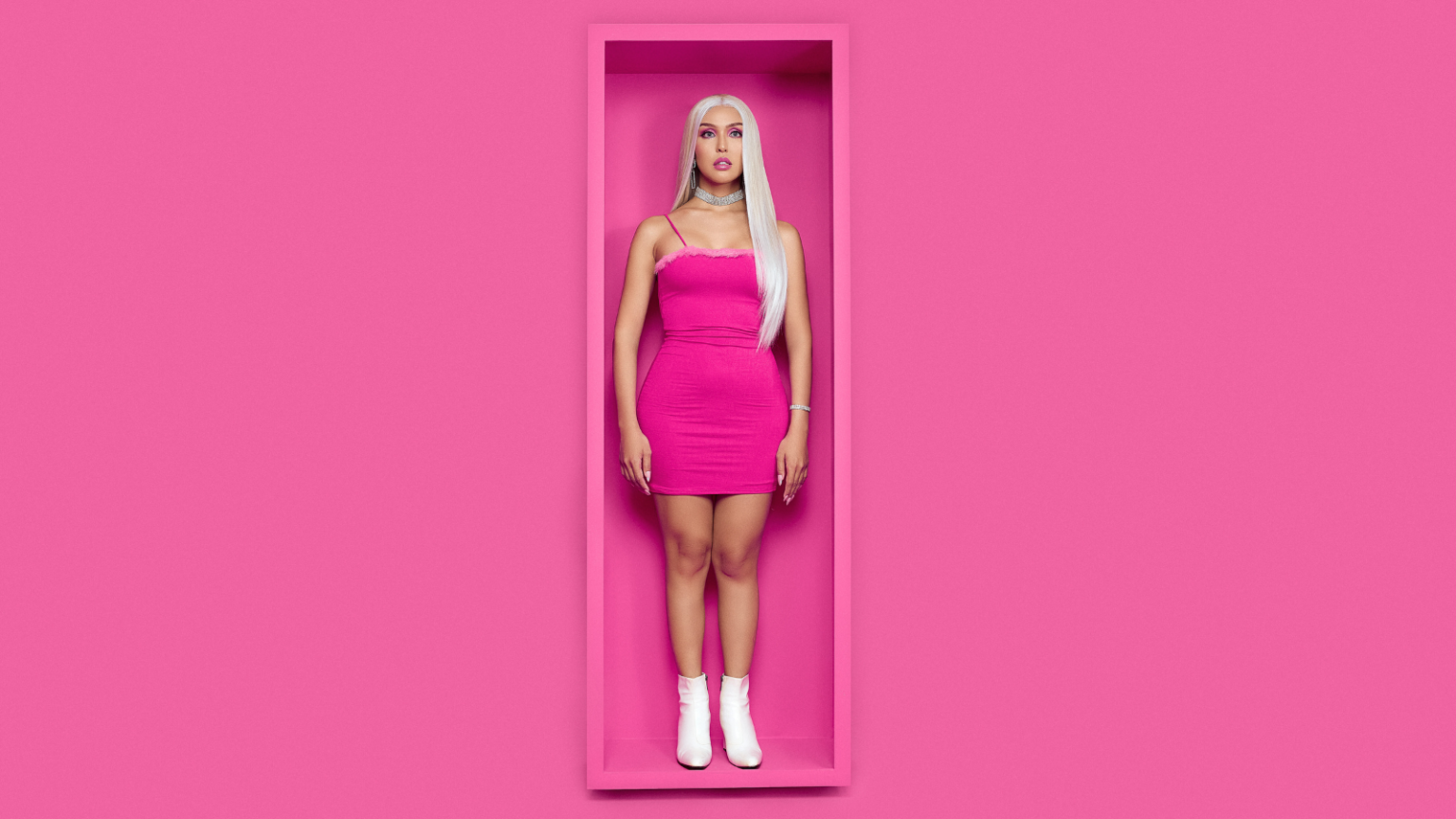 Photo of a thin, blonde feminine person with light skin wearing a hot pink mini dress and standing in a stiff pose inside a large display box